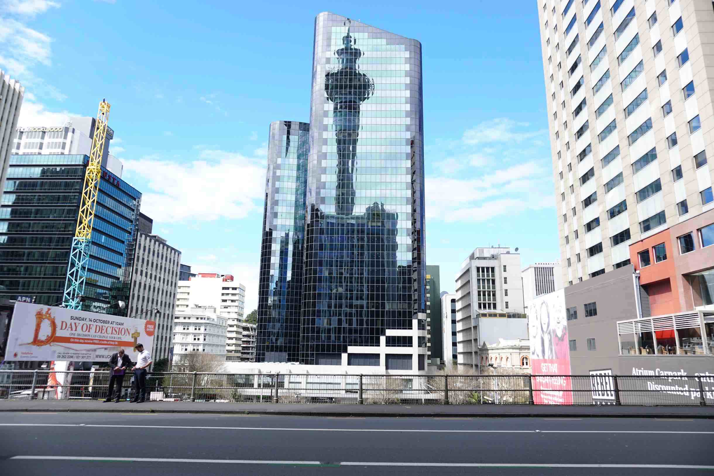 Along a street in downtown Auckland with Sky Tower, the most prominent structure in the city, reflected on an office building.